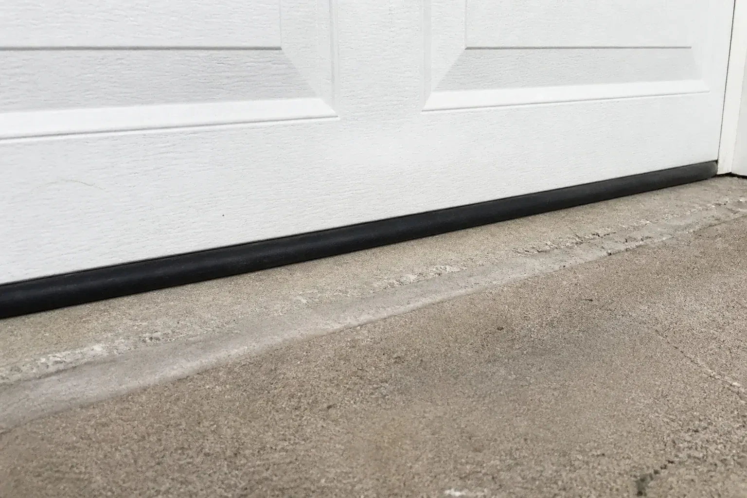 Black bottom seal along a closed white garage door at a concrete threshold. — Garage Door Bottom Seal Replacement, New Tecumseth
