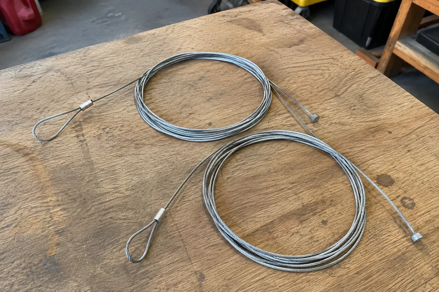 Two coiled steel cables with loop and stop ends on a workbench. — Garage Door Cable Replacement, New Tecumseth