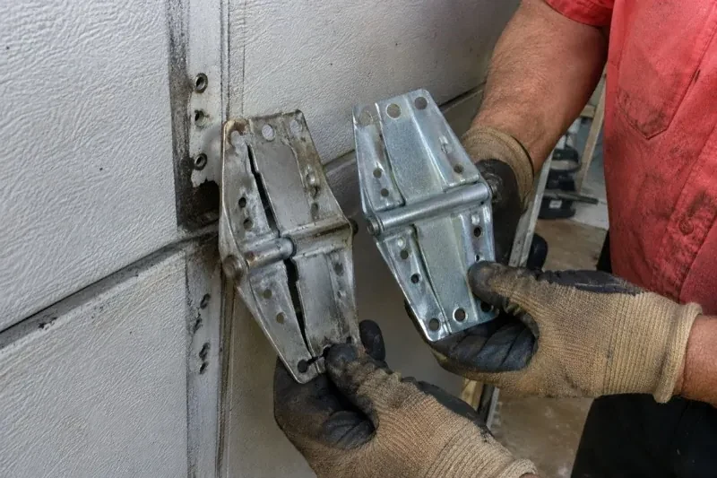 Gloved hands holding two metal garage-door hinges side by side, one discolored with a visible crack and the other bright silver. — Garage Door Hinge Replacement, New Tecumseth