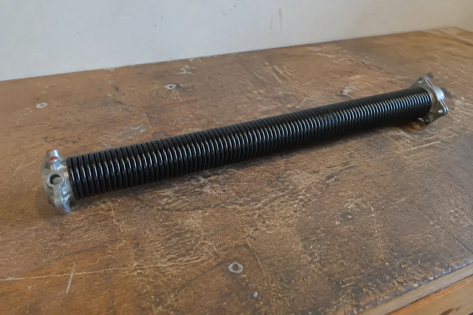 Detached black coil spring with metal end fittings on a workbench. — Torsion Spring Replacement, New Tecumseth